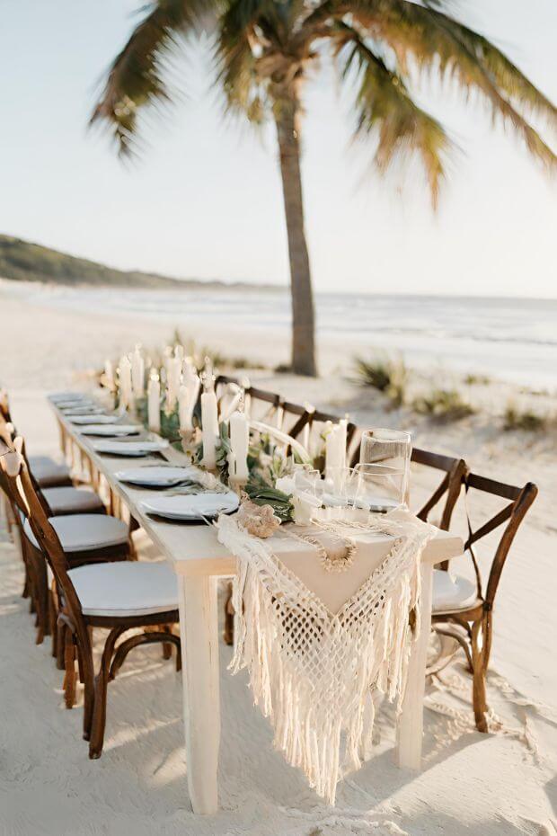 Beach wedding setup with lace table runner, surrounded by wooden chairs and adorned with place settings
