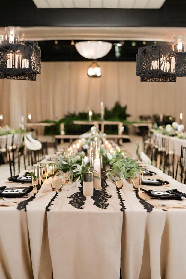 White linen tablecloth with black lace runners and gold candlesticks, adorned with greenery, candles, and silverware