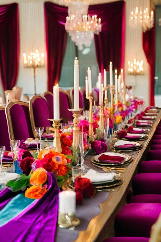 Dining table with runner, gold candlesticks, and vibrant flowers, surrounded by purple drapes