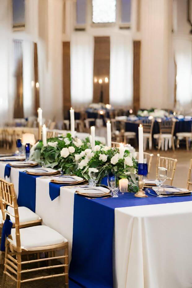 Wedding reception with long dining table set, white linen, gold flatware, and blue and white centerpieces
