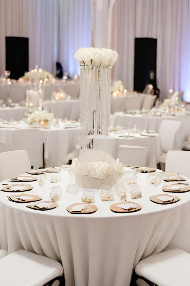 White and gold themed wedding reception with white linens, gold place settings, and tall vase centerpiece with white flowers