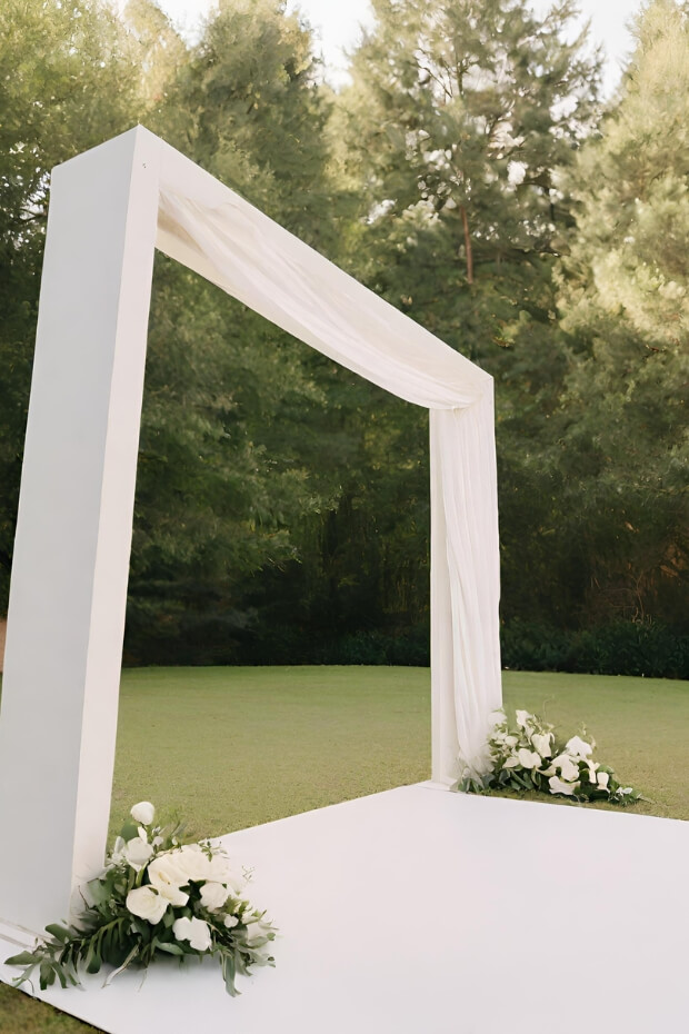 White wedding arch in lush green field with flowers and greenery