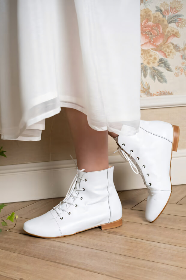 White leather boots with lace-up closure, flat heel, and bohemian aesthetic.